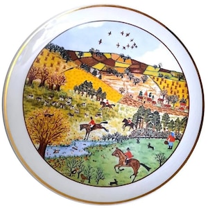 Puede incluir: Un plato de cerámica blanco con un borde dorado presenta una pintura colorida de una escena campestre con jinetes a caballo, un pueblo y una bandada de pájaros volando por encima.