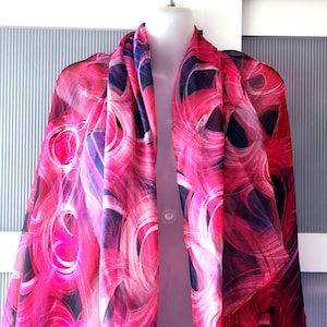 May include: A vibrant pink scarf with a swirling pattern of red, white, and dark blue. The scarf is draped over a mannequin, showcasing its flowing design. The fabric appears lightweight and sheer, perfect for adding a pop of color to any outfit.