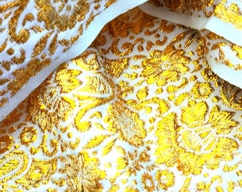 May include: Close-up of a white fabric with a gold floral pattern. The gold design has a metallic sheen, creating a luxurious look. The fabric is folded, revealing a white border.