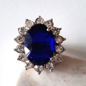 May include: A silver ring featuring a large, oval blue gemstone encircled by smaller, clear gemstones.