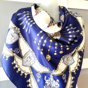 May include: A blue silk scarf with a white and gold pattern of chains, keys, and shields. The scarf is folded and draped over a mannequin's neck.