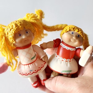 Op de afbeelding: Twee Cabbage Patch Kids poppen met geel garen haar, die rode en witte jurken dragen. Eén pop houdt een roze fles.