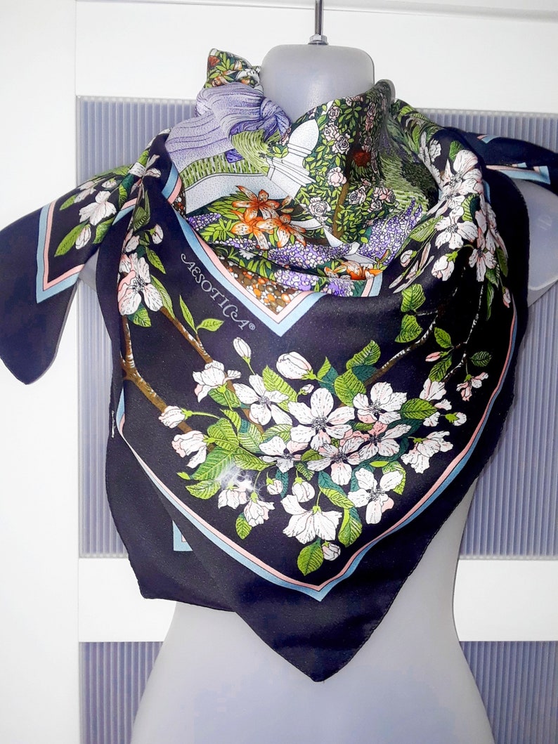 May include: A black silk scarf with a floral print featuring white flowers, green leaves, and a blue and pink border. The scarf has the word "AESTHETICA" printed in white on the border.