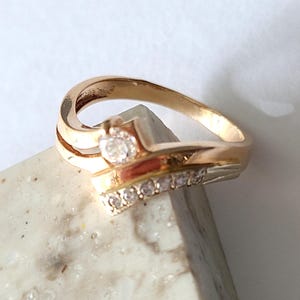 Può includere: Un anello dorato dal design curvo. L'anello presenta una grande gemma trasparente e una fila di gemme più piccole. L'anello è appoggiato su una superficie chiara, mostrando i suoi dettagli intricati e il suo stile elegante.