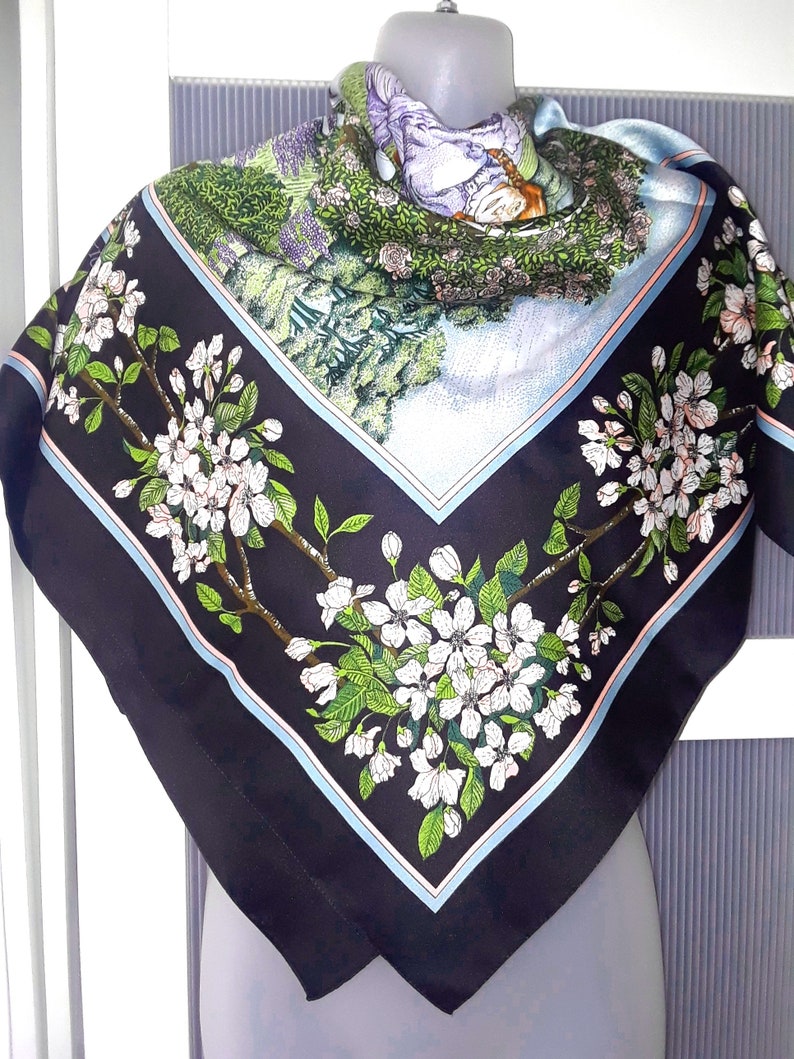 May include: A black silk scarf with a floral print of white blossoms and green leaves. The scarf has a blue and pink border.