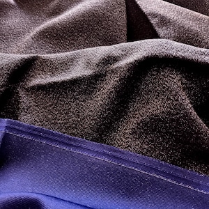 May include: Close-up of draped fabric in shades of dark gray and blue. The top layer is a textured, shimmering gray, while the bottom is a smooth, deep blue with a visible seam. The fabric is folded, creating shadows and highlights.
