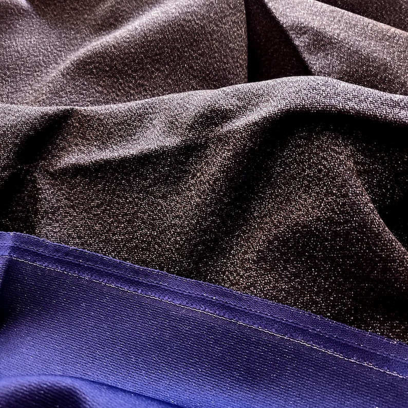 May include: Close-up of draped fabric in shades of dark gray and blue. The top layer is a textured, shimmering gray, while the bottom is a smooth, deep blue with a visible seam. The fabric is folded, creating shadows and highlights.