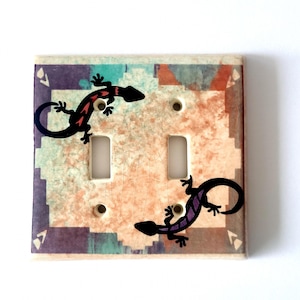 May include: A double light switch cover with a southwestern design featuring two stylized lizards, one red and one purple, on a beige background with turquoise and purple accents.