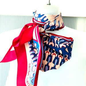 May include: A pink, blue, and red silk scarf with a geometric pattern. The scarf is tied around a mannequin's neck and the brand name "monique matti" is visible on the scarf.