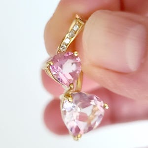 May include: A gold-toned earring featuring two heart-shaped pink gemstones. The top portion of the earring has a small section with clear accent stones. The earring is held up by a person's hand.