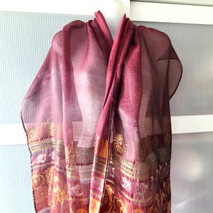 May include: A sheer burgundy scarf with gold floral and geometric patterns along the border. The scarf is draped over a mannequin, showcasing its flowing fabric and design. The scarf's color is a deep, rich shade of red.