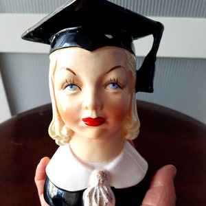 Könnte beinhalten: Eine Keramikbüste einer Frau, die eine schwarze Abschluss-Kappe und -Gown trägt. Die Frau hat blondes Haar, blaue Augen und rote Lippen. Die Büste ist im Vintage-Stil und wäre ein einzigartiges Dekorationsstück.