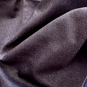 May include: Close-up of a dark purple fabric with a subtle sheen. The material is folded, creating shadows and highlighting the texture. The fabric appears to be soft and drapes naturally, suggesting it could be used for clothing or home decor.