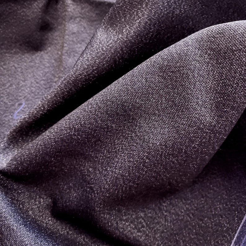 May include: Close-up of a dark purple fabric with a subtle sheen. The material is folded, creating shadows and highlighting the texture. The fabric appears to be soft and drapes naturally, suggesting it could be used for clothing or home decor.