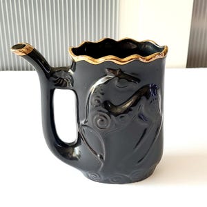 May include: Black ceramic shaving mug with a gold rim and spout. The mug features an embossed design of a horse. The handle has a rectangular opening. A decorative item for a bathroom.
