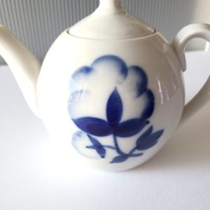 Puede incluir: Tetera blanca con un diseño floral azul. La tetera tiene un cuerpo redondo y una tapa.