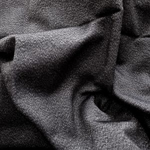 May include: Close-up of a dark gray fabric with a textured surface. The fabric is heavily draped, creating deep shadows and highlights. The material appears to be woven, with a subtle sheen. The overall effect is one of depth and richness.