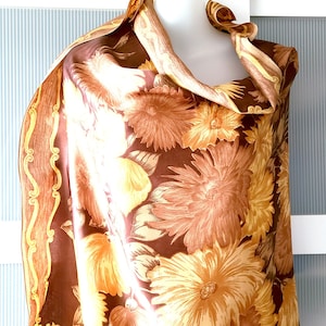 May include: A brown and yellow floral scarf draped over a mannequin. The scarf features large, stylized flowers in shades of brown, yellow, and orange. The edges of the scarf have a decorative border. The scarf is made of a silky material.