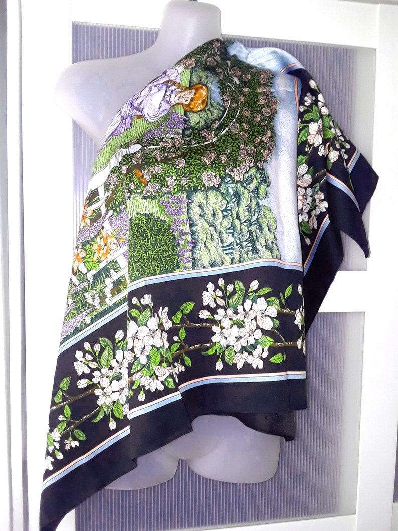 May include: A black and white silk scarf with a floral design. The scarf features a woman sitting in a garden with flowers and trees. The scarf has a blue, white, and pink border.