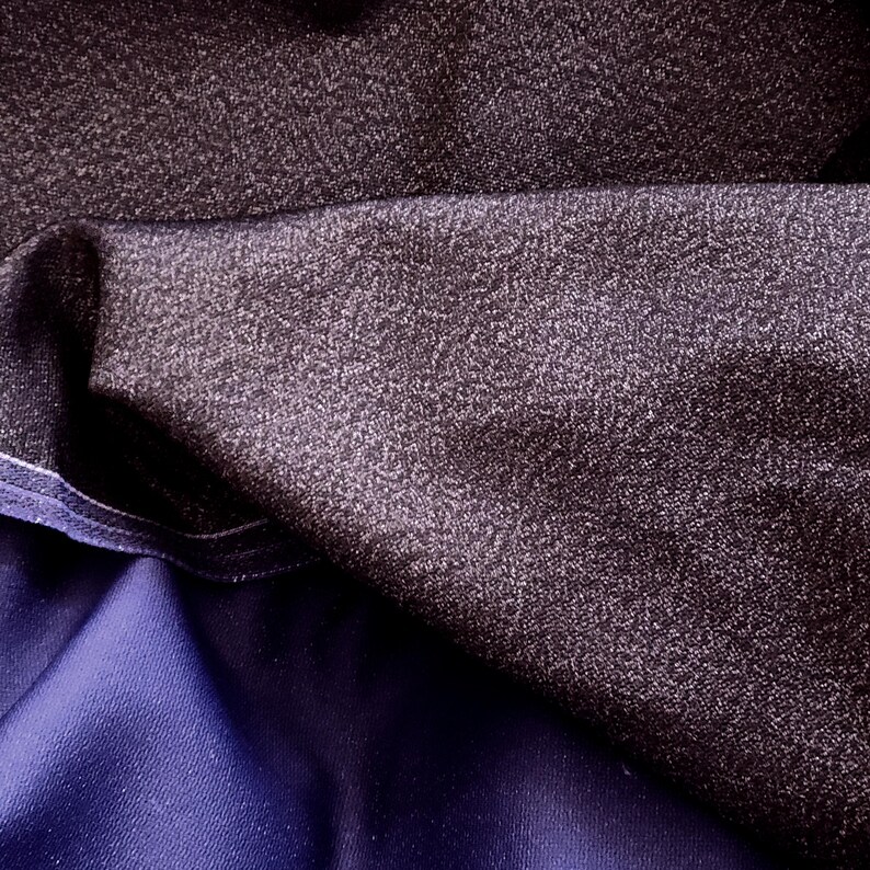 May include: Close-up of dark fabric with a textured, speckled appearance. The fabric is a deep charcoal color with a subtle sheen, and it is layered over a smooth, dark blue material. The textures and colors create a visually interesting contrast.