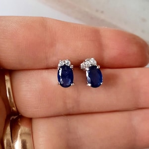 May include: A pair of silver earrings featuring oval-shaped blue sapphire gemstones with smaller clear gemstones around the edge.