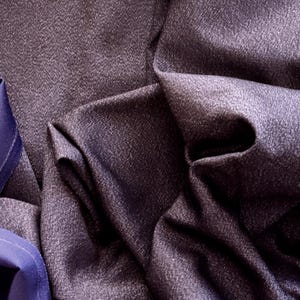 May include: Close-up of a dark purple fabric with a subtle textured pattern. The fabric is draped, creating folds and shadows that highlight its texture. A section of blue fabric is visible on the left side, adding contrast.