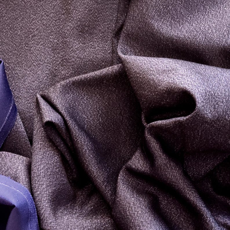 May include: Close-up of a dark purple fabric with a subtle textured pattern. The fabric is draped, creating folds and shadows that highlight its texture. A section of blue fabric is visible on the left side, adding contrast.