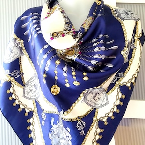 Op de afbeelding: Een blauwe en witte zijden sjaal met een gouden kettingpatroon en een kroonontwerp. De sjaal is gevouwen en gedrapeerd over de nek van een mannequin.