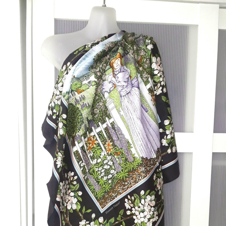 May include: A black silk scarf with a colorful floral print featuring a woman standing in a garden. The scarf has a blue and white border and the text "Elizabeth Parker" in the bottom right corner.
