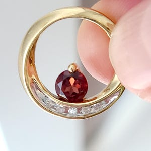 May include: A gold pendant with a circular design. The pendant features a large, round, deep red gemstone suspended within the circle. The gold circle is also adorned with small, clear gemstones. The pendant is held by a finger.