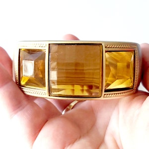 May include: Gold-coloured metal cuff bracelet with three large, faceted amber-coloured rectangular stones. The bracelet has a textured rope-like design along the edges, creating a vintage aesthetic. The bracelet is a statement piece.