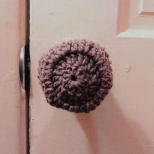May include: A circular, brown, crocheted cover made of yarn, designed to fit over a round door knob. The texture is created by dense, concentric stitches that radiate from the center, providing a soft, thick layer over the knob surface.