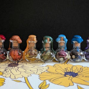 May include: Seven small, clear glass bottles with cork stoppers and colorful wax seals. Each bottle contains dried herbs and flowers. The wax seals are red, brown, orange, green, blue, and purple. The bottles are arranged on a floral patterned surface.