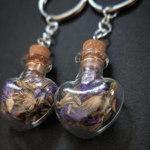 May include: Two heart-shaped glass bottles filled with dried flowers and sealed with cork stoppers. The bottles are attached to silver keychains. The flowers inside are purple and tan. The background is black.
