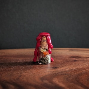 May include: A small glass bottle with a cork stopper, filled with dried herbs and flowers. The bottle is sealed with bright red wax that has dripped down the sides. The bottle sits on a wooden surface.