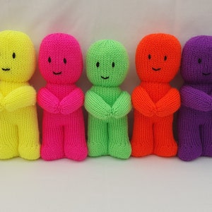 Op de afbeelding: Zeven gebreide poppen op een rij, elk in een andere heldere kleur: rood, geel, roze, groen, oranje, paars en blauw. Elke pop heeft een eenvoudig gezicht met een glimlach en gekruiste armen.