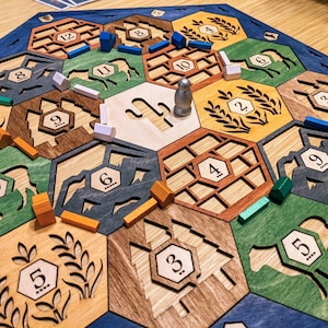 May include: A wooden board game with hexagonal tiles in various colors, including green, brown, and blue. The game board features wooden pieces, including houses, roads, and a silver robber piece. The game is set up and ready to play.