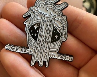 May include: Black and white enamel pin featuring a stylized astronaut helmet with a cloud and rain design. The pin includes the text "COME BACK TO EARTH" in a black font. The pin is a unique accessory.