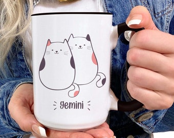 May include: White ceramic mug with a black handle. The mug features a cartoon illustration of two white cats with pink and black markings. The text "gemini" is printed below the cats.