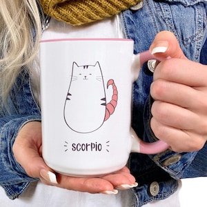 May include: White ceramic mug with a pink rim and handle. The mug features a cartoon cat with a scorpion tail and the text "Scorpio".