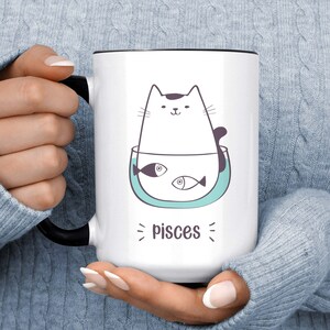 May include: White ceramic mug with a black rim and handle. The mug features a cartoon illustration of a white cat with black ears and a tail, sitting in a blue bowl with three fish. The text "Pisces" is written below the cat.