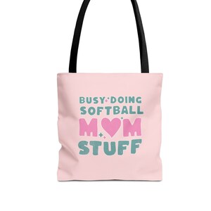 May include: Pink tote bag with black handles. The bag has a graphic that says "Busy Doing Softball Mom Stuff" with a pink heart in the middle of the word "Mom".
