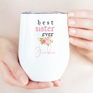 Puede incluir: Un vaso de vino de acero inoxidable blanco con un diseño floral y el texto "best sister ever" y el nombre "Jackie" en cursiva.