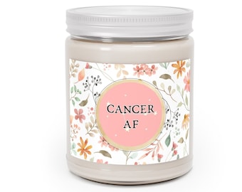 Könnte beinhalten: Ein weißes Kerzengefäß mit einem silbernen Deckel und einem Etikett mit Blumenmuster. Das Etikett hat einen rosa Kreis mit dem Text "CANCER AF" in Schwarz.