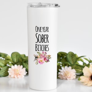May include: White stainless steel tumbler with a black floral design and the text "One Year Sober Bitches".