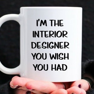 May include: White ceramic mug with the text "I'M THE INTERIOR DESIGNER YOU WISH YOU HAD" in black. The mug is held by a person with dark nail polish.