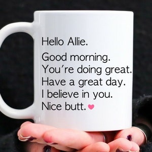May include: White ceramic mug with a black handle, featuring the text "Hello Allie. Good morning. You're doing great. Have a great day. I believe in you. Nice butt." in black font. A pink heart is at the end of the text.