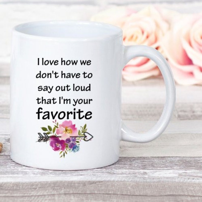 May include: White ceramic mug with a black and white quote that reads "I love how we don't have to say out loud that I'm your favorite". The quote is surrounded by a floral design with pink, purple, and green flowers.