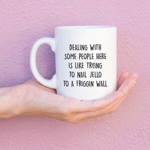 Może przedstawiać: Biały ceramiczny kubek z zakrzywionym uchwytem, trzymany w dłoni. Kubek ma czarny tekst, kt&oacute;ry brzmi: "DEALING WITH SOME PEOPLE HERE IS LIKE TRYING TO NAIL JELLO TO A FRIGGIN WALL."