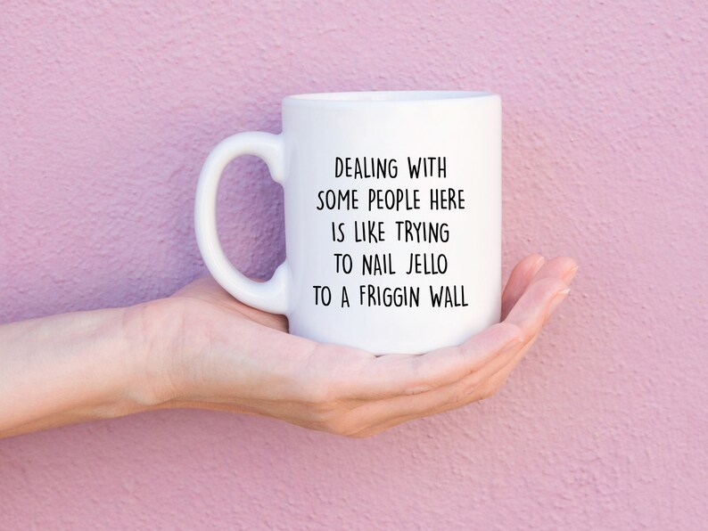 Może przedstawiać: Biały ceramiczny kubek z zakrzywionym uchwytem, trzymany w dłoni. Kubek ma czarny tekst, kt&oacute;ry brzmi: "DEALING WITH SOME PEOPLE HERE IS LIKE TRYING TO NAIL JELLO TO A FRIGGIN WALL."